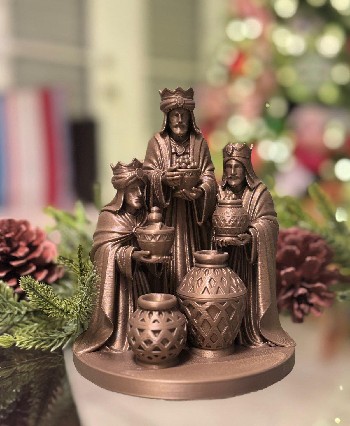 Escultura decorativa de los Tres Reyes Magos en tono bronce, con coronas, vestimentas detalladas y ofrendas sobre una base circular, frente a un fondo navideño con luces desenfocadas.